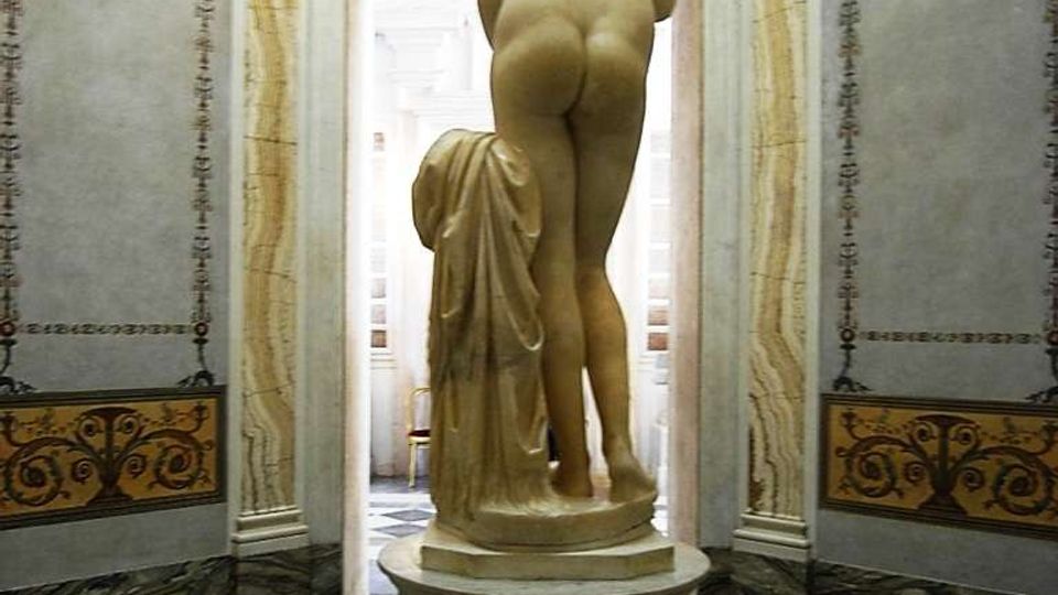 Interno di una sala museale classica con pavimento in marmo geometrico e pareti decorate, inquadrata verso una porta luminosa. Al centro, su un piedistallo, è esposta una statua in marmo a figura nuda vista di spalle, con drappo scolpito accanto.