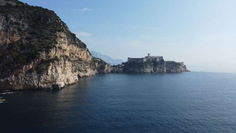 Veduta costiera con alte scogliere rocciose a picco sul mare calmo; in lontananza si vede un promontorio con una fortezza o complesso di edifici storici affacciati sull’acqua. Cielo sereno e costa frastagliata che prosegue verso l’orizzonte.