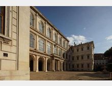 Cortile interno di un palazzo storico in pietra, con loggiato ad archi al piano terra e grandi finestre ad arco sui livelli superiori. Lo spazio è ampio e pavimentato, circondato da facciate classiche sotto un cielo sereno.
