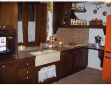 Cucina domestica con mobili in legno scuro a L, piano di lavoro con lavello e rubinetto in rame e alzatina piastrellata. Si vedono finestra con tende, forno incassato, mensole con barattoli e utensili, pavimento in piastrelle e un frigorifero arancione sulla destra.