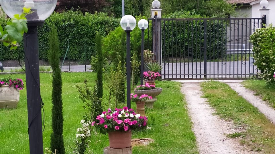 Vialetto ghiaioso in un giardino con prato, vasi di fiori e lampioni con sfere trasparenti, che conduce a un cancello metallico. Sullo sfondo si vedono alberi fitti su un pendio e alcune case con tetti in tegole.