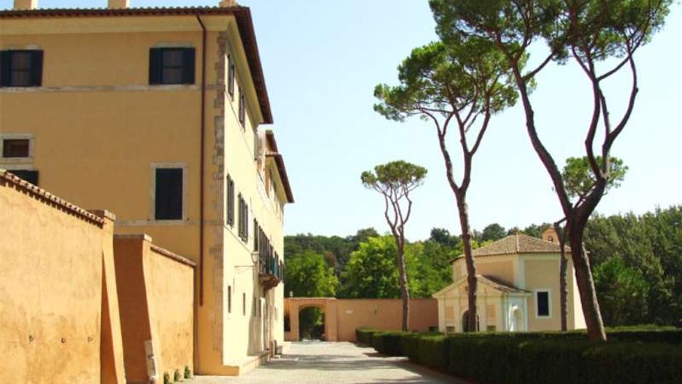 Viale con pavimentazione in ghiaia accanto a un grande edificio storico color ocra con persiane scure e tetto in coppi. Sul lato opposto ci sono siepi potate e alti pini marittimi, con un piccolo edificio e un arco d’ingresso sullo sfondo.