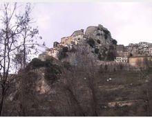 Borgo storico arroccato su una rupe rocciosa, con case in pietra addossate al pendio e resti di una fortificazione in cima. In primo piano si vedono alberi spogli e terreni collinari sotto un cielo nuvoloso.