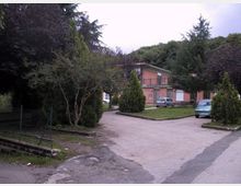 Strada asfaltata interna con parcheggio e aiuole alberate, circondata da alberi alti e vegetazione fitta. Sullo sfondo si vede un edificio basso in mattoni rosa con balconi e tetto spiovente, con alcune auto parcheggiate.