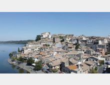 Vista panoramica di un borgo collinare con case in pietra dai tetti in coppi, addossate l’una all’altra, e una chiesa in posizione elevata. Il paese si affaccia su un lago con riva alberata e strada lungo l’acqua sotto un cielo sereno.