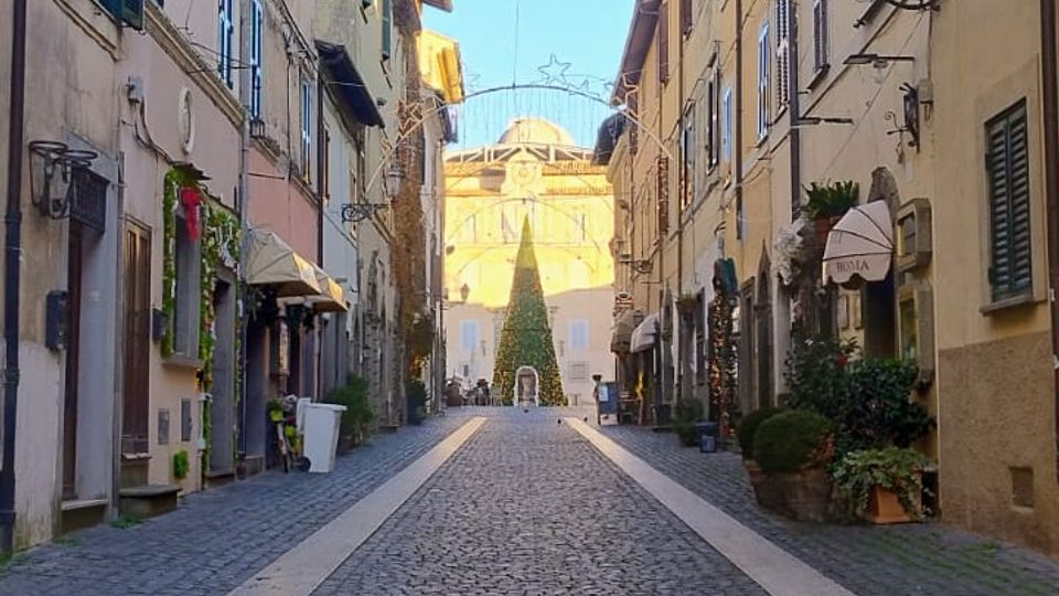 Strada stretta in un centro storico con pavimentazione in sampietrini, fiancheggiata da edifici a più piani con persiane e lampioni a parete. In alto sono appese luminarie a forma di stella e, in fondo alla via, si vede una piazza con un grande edificio e un albero decorato.