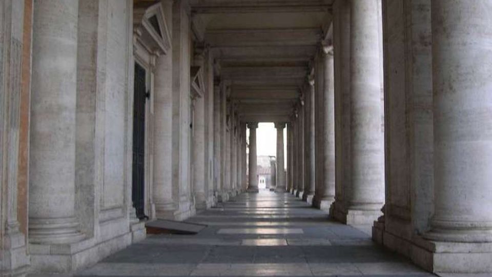 Lungo portico in pietra con alte colonne cilindriche e soffitto a travi, visto in prospettiva verso un’apertura luminosa sul fondo. Pavimento a grandi lastre grigie e pareti con ingressi incorniciati lungo il lato sinistro.