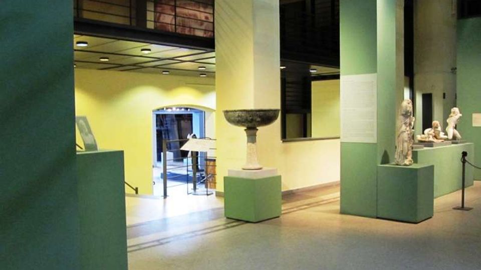 Interno di un museo o spazio espositivo con grandi pilastri e pareti verde chiaro, pavimento in piastrelle e illuminazione a soffitto. Su piedistalli sono esposte sculture e un grande bacile in pietra, con una passerella e un soppalco visibili sullo sfondo.