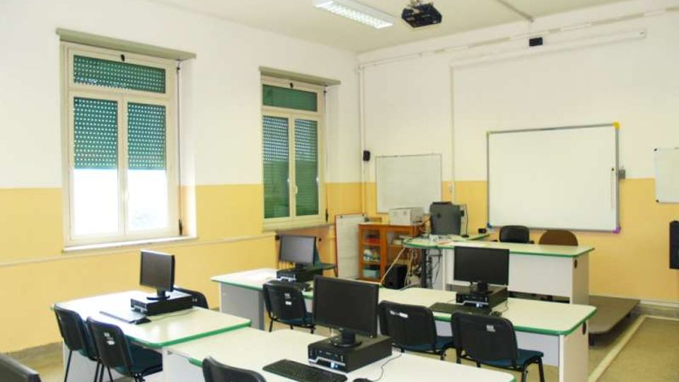 Aula informatica con banchi disposti in file, postazioni con monitor e sedie nere, pareti bianche e gialle e pavimento grigio. Sul fondo ci sono una cattedra, una lavagna bianca e un proiettore a soffitto, con finestre laterali dotate di tapparelle verdi.