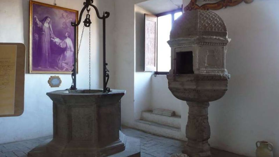Interno di una piccola cappella o museo con pareti bianche e pavimento in pietra, con un pozzo in pietra dotato di struttura in ferro al centro. A destra c’è un fonte battesimale scolpito con cupola decorata e una finestra aperta che illumina l’ambiente; sulla parete si vede un grande dipinto religioso incorniciato.