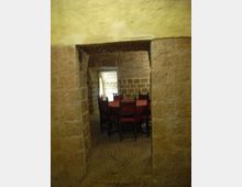 Passaggio ad arco in pietra con soffitto a volta in mattoni, che conduce a una sala con pareti in blocchi di pietra. All’interno si vedono tavoli con tovaglie rosse e sedie in legno, con luce naturale proveniente da una finestra sul fondo.