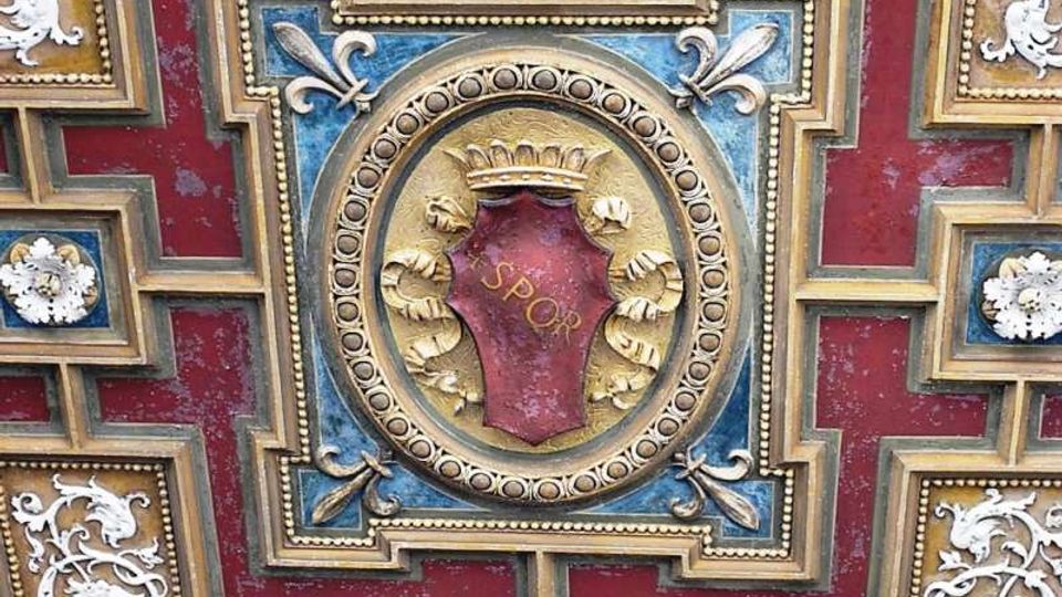 Dettaglio ravvicinato di una decorazione architettonica a soffitto con motivo geometrico rosso e blu, cornici dorate e ornamenti in stucco bianco. Al centro, un medaglione circolare con stemma dorato e la scritta “SPQR” su fondo rosso.