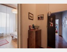 Corridoio interno di un appartamento con pavimento in legno, un mobile basso in legno con oggetti decorativi e quadri alle pareti, affiancato da un armadio scuro. A sinistra si apre una stanza luminosa con grandi tende bianche e lampada a sospensione, mentre a destra il corridoio prosegue verso altre stanze.