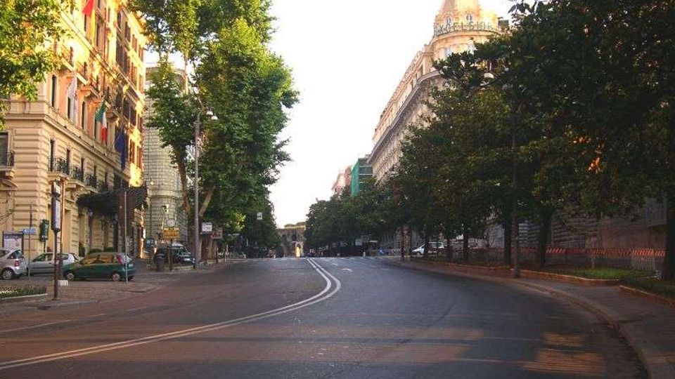 Ampio viale urbano curvo con doppia linea centrale, fiancheggiato da alberi e marciapiedi. Ai lati si vedono palazzi storici con facciate ornate e, sullo sfondo, un grande edificio con cupola.