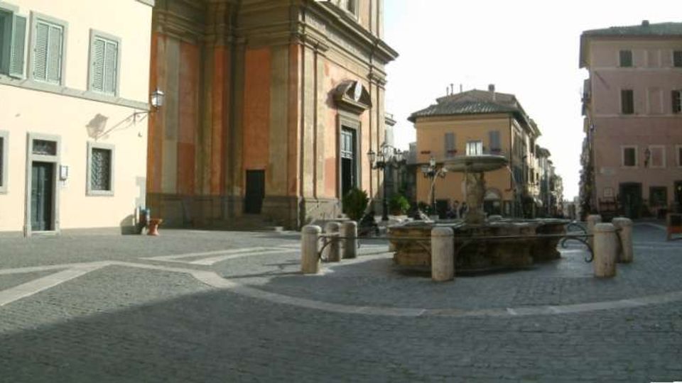 Piazza lastricata in pietra con una fontana circolare al centro e paletti dissuasori intorno. Sullo sfondo si vedono edifici storici dai toni chiari e la facciata laterale di una chiesa con alti pilastri e dettagli in pietra.