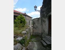 Vicolo stretto in un borgo con muri in pietra e intonaco, gradini in cemento e una lanterna in ferro battuto fissata alla parete. Ai lati si vedono case con tetti in tegole rosse, vegetazione spontanea e cielo azzurro sullo sfondo.