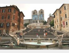 Ampia scalinata monumentale in una piazza urbana, con fontana circolare in primo piano e edifici storici ai lati. In cima alla scalinata si vede la facciata di una chiesa in restauro con impalcature e teli decorati.