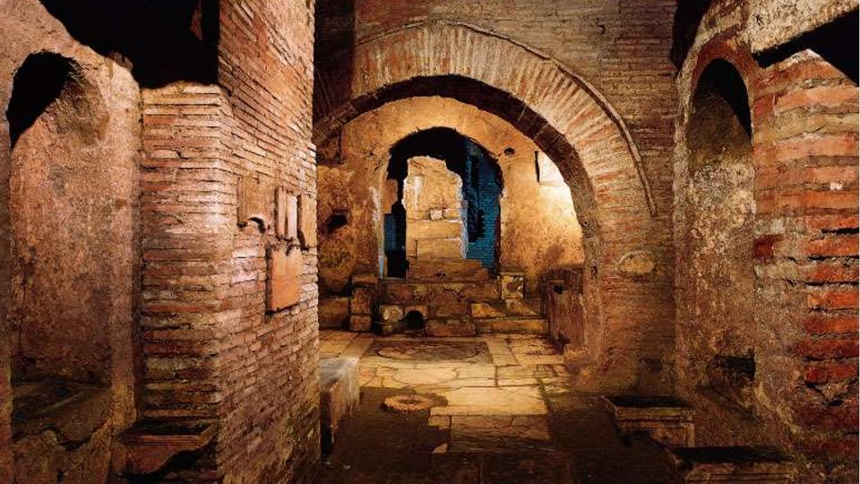 Interno di rovine sotterranee in mattoni e pietra, con volte e archi che conducono a un passaggio centrale. Pavimento in lastre irregolari e pareti antiche illuminate da luce calda, con nicchie e gradini sullo sfondo.