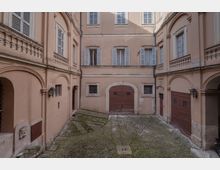 Cortile interno di un edificio storico con facciate color beige, archi e finestre con persiane. Al centro si vede un grande portone in legno e una pavimentazione in ciottoli con motivi geometrici.