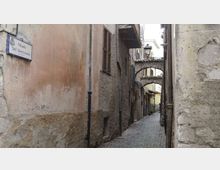 Vicolo stretto con pavimentazione in ciottoli tra edifici in pietra e intonaco deteriorato, con finestre dotate di persiane e tubi di scolo a vista. In fondo si vedono un arco di collegamento tra gli edifici e un lampione, con una targa stradale sul muro a sinistra.