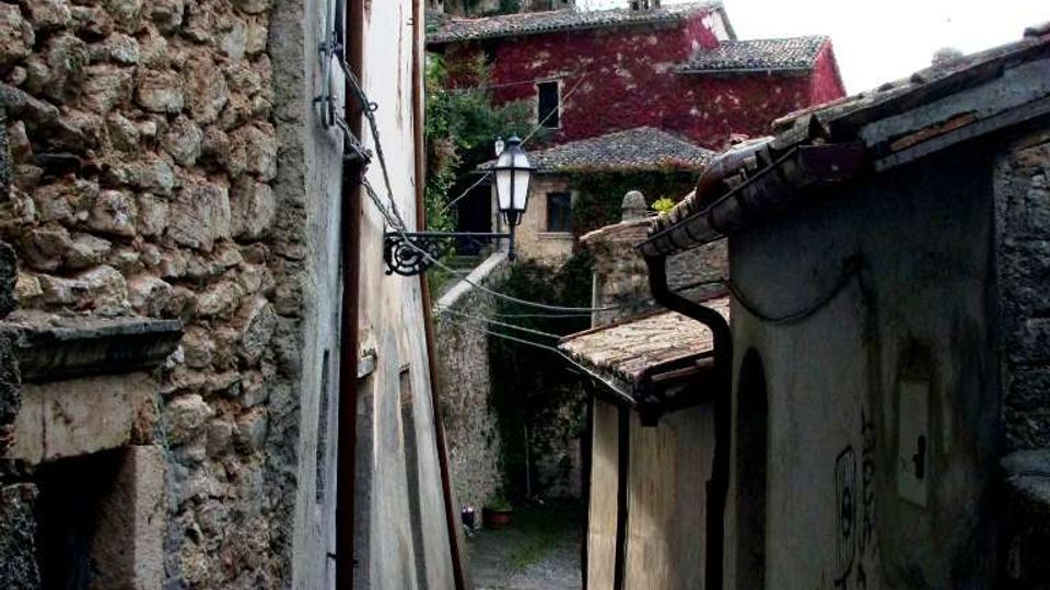 Vicolo stretto in un borgo antico, con pavimentazione in pietra e muri alti in pietra e intonaco ai lati. Una lanterna a parete e cavi sospesi attraversano il passaggio, con case dai tetti in tegole visibili sullo sfondo in salita.