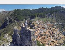 Veduta aerea di un borgo montano con case addossate dai tetti in tegole e strade strette, ai piedi di una rupe rocciosa. In primo piano, una fortificazione in pietra con statua sulla sommità domina il paese, circondato da pendii boscosi e montagne sullo sfondo.