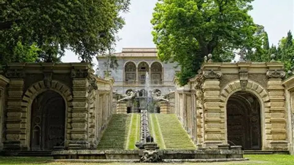 Giardino formale con una lunga scalinata in pietra e un canale d’acqua centrale che sale verso una villa storica con loggia ad archi. Ai lati della scalinata ci sono due grandi portali in muratura decorata e alberi ad alto fusto che incorniciano la scena.