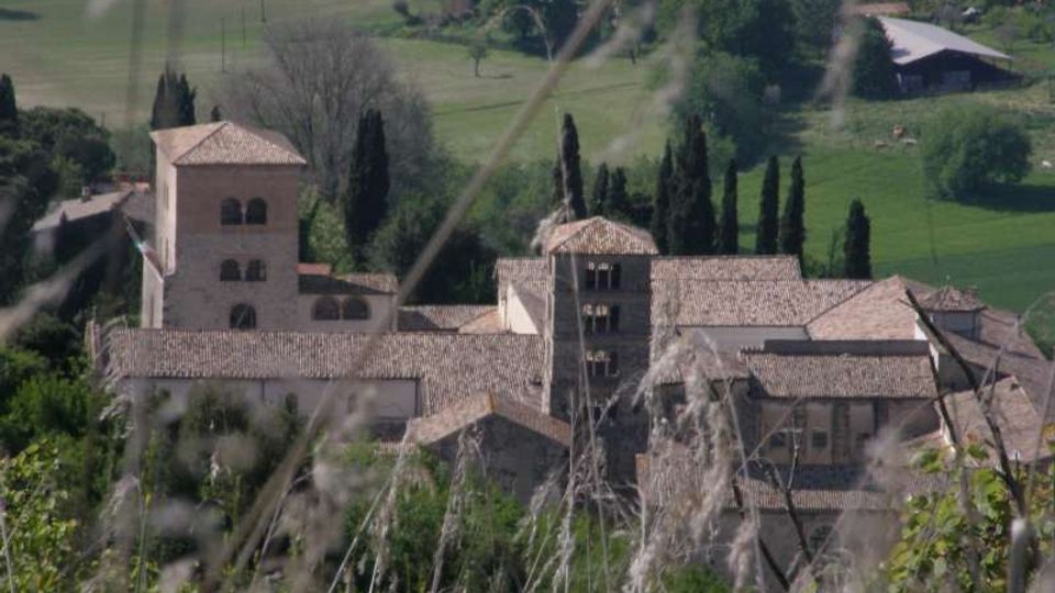 Complesso di edifici in pietra con tetti in coppi e due torri quadrate, immerso in una campagna collinare con campi verdi e filari di cipressi. In primo piano si vedono erbe e rami sfocati che incorniciano la vista dall’alto.