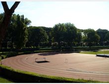 Pista di atletica all’aperto con curve marcate e alcune barriere per l’allenamento, circondata da siepi e da un’area verde. Sullo sfondo si vede una fila di alberi alti che delimita il parco sotto un cielo sereno.