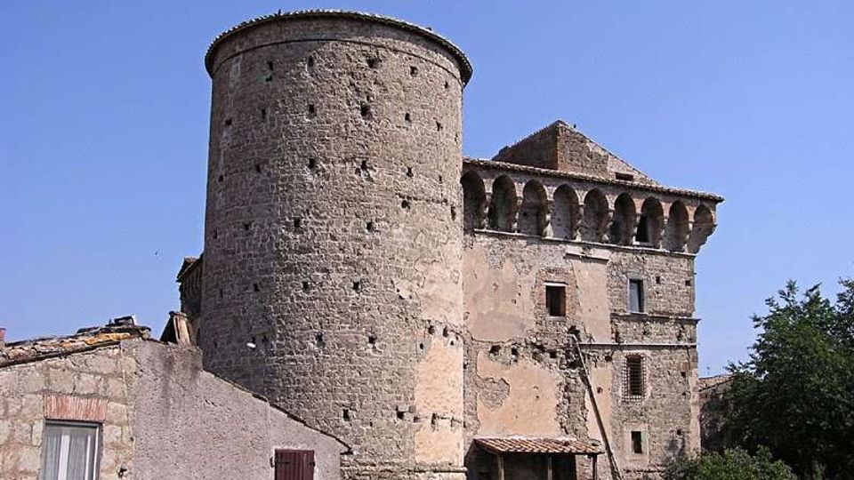 Antico edificio fortificato in pietra con grande torre cilindrica e struttura adiacente con loggiato ad archi, affacciato su un piccolo nucleo di case. Sullo sfondo si vede il cielo sereno e vegetazione ai margini dell’abitato.