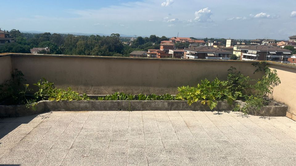 Terrazza panoramica con pavimento piastrellato e muretto perimetrale, con una lunga fioriera in cemento con vegetazione. Sullo sfondo si vede un quartiere di case e alberi, con colline lontane sotto un cielo azzurro con nuvole.