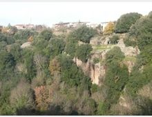 Veduta di una gola boschiva con pareti di roccia e vegetazione fitta, con alberi dai colori autunnali. In alto si vedono alcune case e costruzioni sul bordo della scarpata.