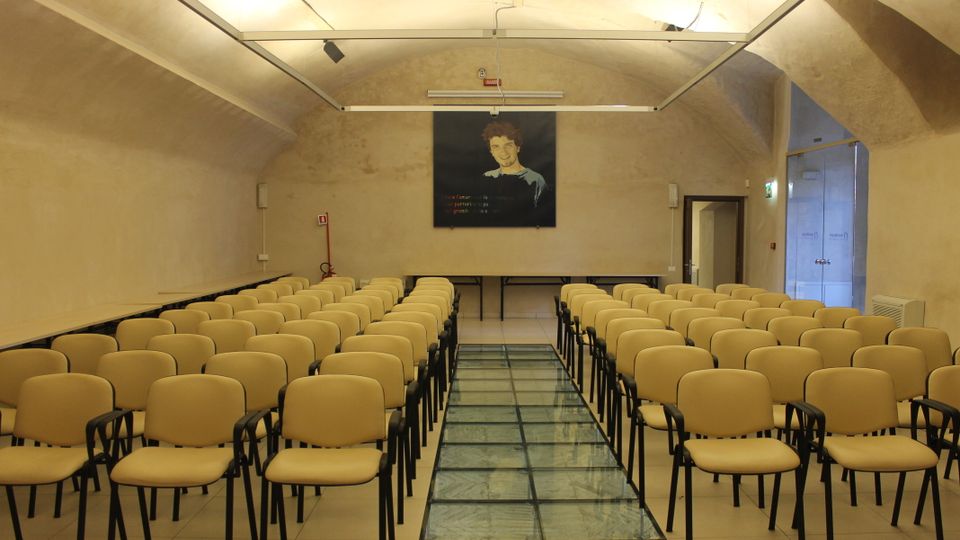 Sala conferenze con soffitto a volta e pareti in intonaco chiaro, illuminata da faretti su binari. File di sedie beige disposte ai lati di un corridoio centrale con pavimento in vetro, con un tavolo e un grande pannello appeso sulla parete di fondo.