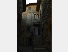 Vicolo stretto tra edifici in pietra con scala in muratura che sale verso un portone ad arco con tenda. Facciate rustiche con persiane e finestre ad arco, grondaie a vista e tetto con tegole.