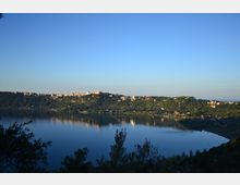 Vista panoramica di un lago calmo con riflessi sull’acqua, circondato da colline boscose. Sulla cresta delle colline si vedono abitazioni e piccoli edifici, sotto un ampio cielo sereno.