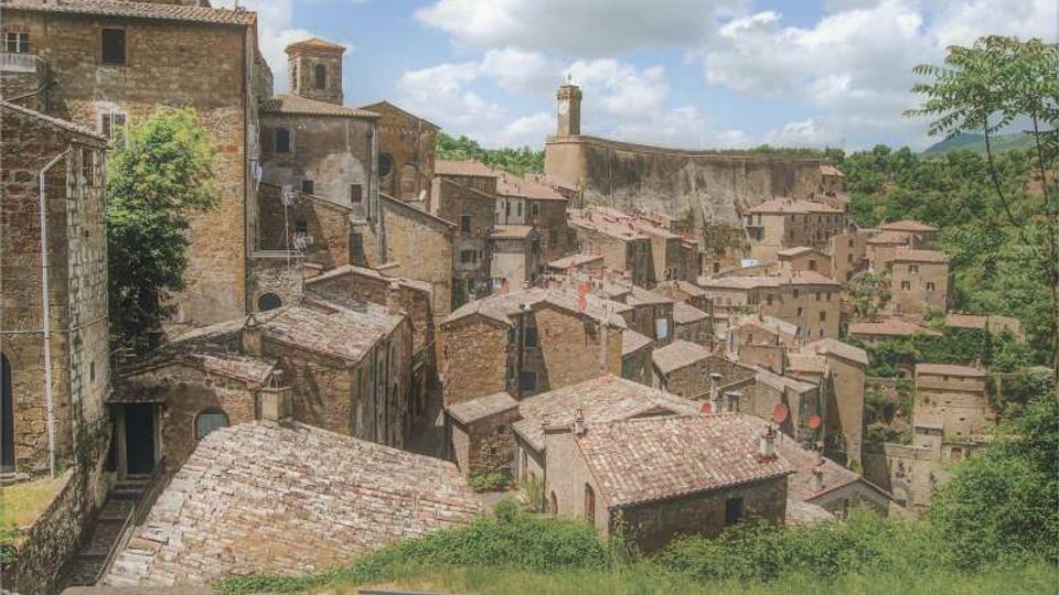 Vista panoramica di un borgo medievale su un pendio, con case in pietra addossate tra loro e tetti in coppi. Sullo sfondo si vedono una chiesa con campanile e una lunga muraglia sopra un’area verde collinare.