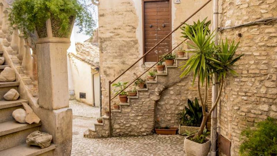 Vicolo acciottolato in un borgo con edifici in pietra e intonaco, una piccola scalinata esterna con ringhiera metallica che sale a una porta in legno. Vasi di terracotta con piante lungo i gradini e un albero in vaso accanto a un muro in pietra.
