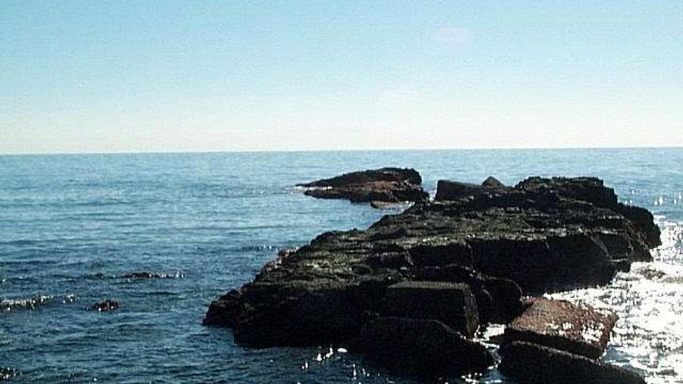 Scogliera rocciosa che si estende nel mare, con grandi massi scuri e acqua calma che riflette la luce. All’orizzonte si vede il mare aperto sotto un cielo sereno e limpido.