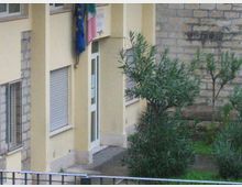 Facciata di un edificio pubblico color giallo chiaro con ingresso, finestre con tapparelle e una bandiera dell’Unione Europea e italiana vicino alla porta. In primo piano si vede un piccolo giardino con arbusti e un muretto di pietra sul lato destro.