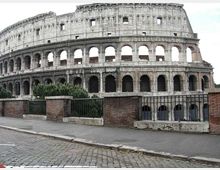 Veduta esterna del Colosseo, anfiteatro romano in pietra con più ordini di archi sovrapposti e aperture. In primo piano si vedono un marciapiede con pavé, una ringhiera metallica e muretti in mattoni con vegetazione bassa.