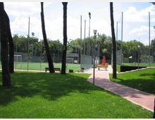 Parco sportivo con campi da calcio in erba sintetica recintati da alte reti e pali di illuminazione. In primo piano un prato con alberi e una passerella pavimentata con panchine e un ombrellone chiuso, circondati da vegetazione.