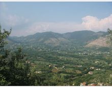 Veduta panoramica di una valle rurale con campi e vegetazione fitta, punteggiata da case sparse, con catene montuose sullo sfondo. In primo piano si vedono alberi e cespugli ai margini dell’inquadratura sotto un cielo parzialmente nuvoloso.