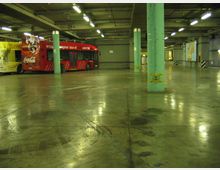 Ampio garage sotterraneo con pavimento in cemento lucido e segni di pneumatici, illuminato da file di lampade al neon e sostenuto da colonne verdi. Sul lato sinistro è parcheggiato un autobus rosso accanto a pareti grigie e cartelloni pubblicitari.
