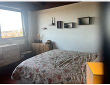Camera da letto luminosa con letto matrimoniale e copriletto floreale, parete bianca con mensole a cubo e soffitto con travi scure. Una grande finestra con tenda grigia illumina la stanza, con cassettiera e mobile decorato vicino alla parete.