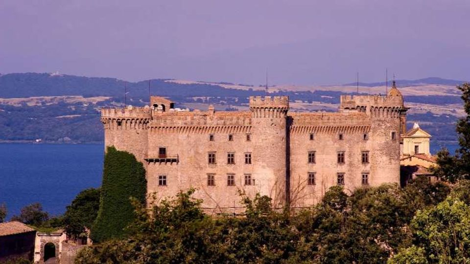 Grande castello in pietra con mura merlate e torri angolari, circondato da vegetazione in primo piano. Sullo sfondo si vede un lago e colline lontane sotto un cielo limpido.