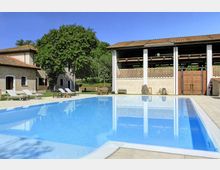 Piscina rettangolare all’aperto con acqua azzurra e bordo in pietra, circondata da lettini e prato. Sullo sfondo si vedono edifici rustici con tetti in coppi e un grande portico aperto con colonne bianche, in un contesto di campagna con alberi.