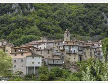 Borgo collinare con case in pietra e intonaco addossate al pendio, tetti in coppi e una piccola torre campanaria al centro. Sullo sfondo si vede un fitto bosco montano con affioramenti rocciosi.