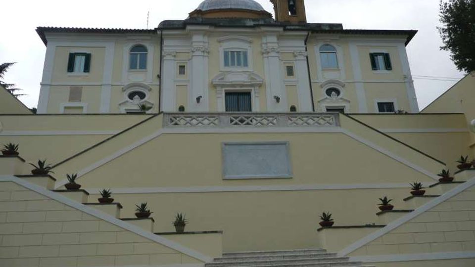Facciata di un edificio storico con elementi barocchi, cupola e campanile, vista dal basso. Un’ampia scalinata centrale in pietra con rampe laterali e vasi di piante conduce a una terrazza davanti all’ingresso.