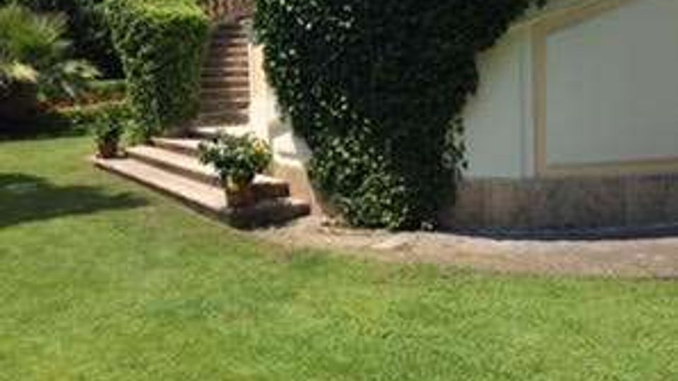 Giardino curato con ampio prato verde e una scalinata in pietra che sale tra siepi potate. Sul lato destro si vede un muro chiaro con base in pietra, affiancato da grandi cespugli arrotondati e vegetazione ornamentale.