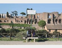 Panchina in un parco con staccionata in legno e cartello “ROMOLO E REMO” in primo piano, con vista su rovine archeologiche in muratura e archi sullo sfondo. Attorno si vedono alberi alti e vegetazione, con un tratto di strada e edifici bassi ai piedi delle rovine.